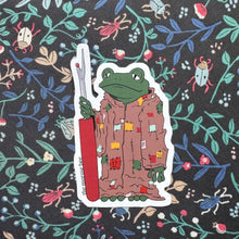 Load image into Gallery viewer, Sticker of a frog character holding a seam ripper on a floral patterned background
